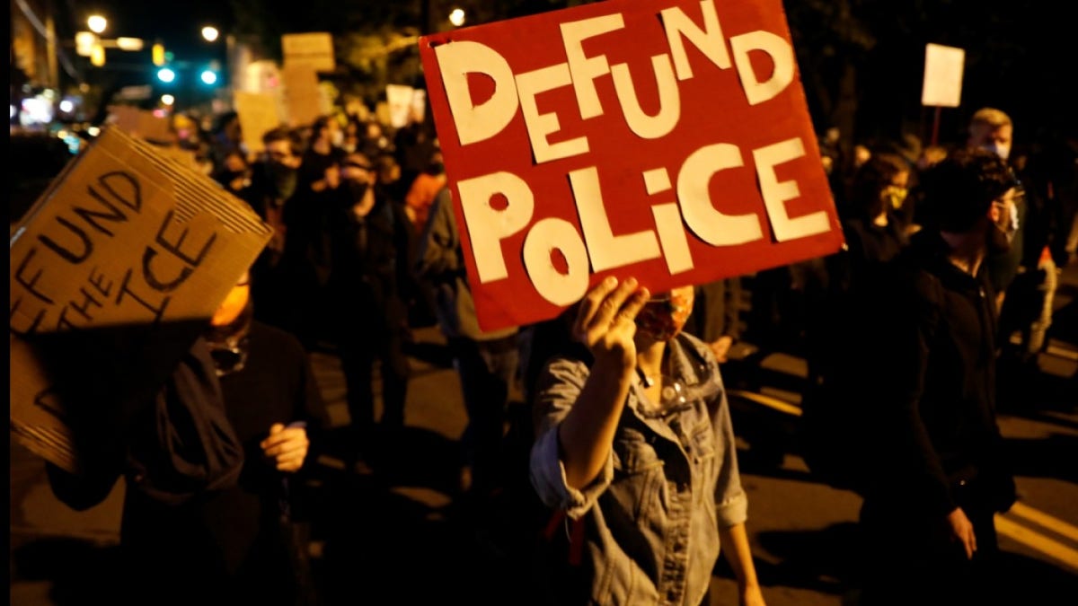 Photo shows a person holding a sign reading "Defund Police" at a protest in 2020